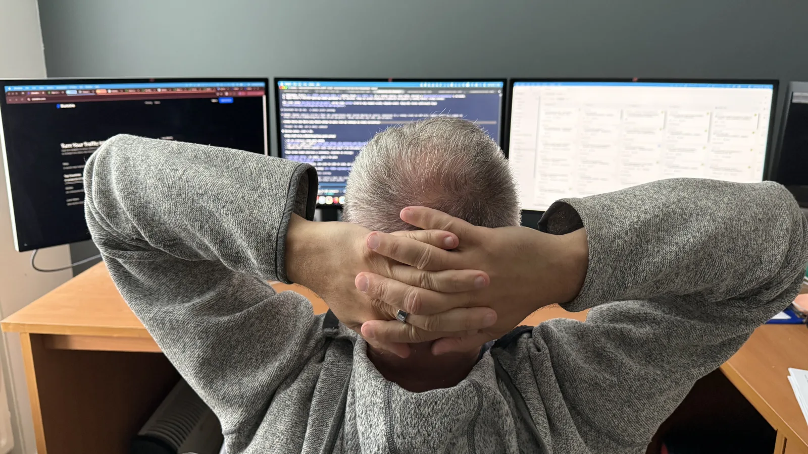 Me at the desk, hands behind my head, three monitors doing the work